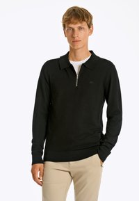 Maglione polo nero con zip e maniche lunghe, polsini e orlo a coste, realizzato in tessuto morbido; indossato sopra una camicia bianca. Dettaglio del logo minimalista.