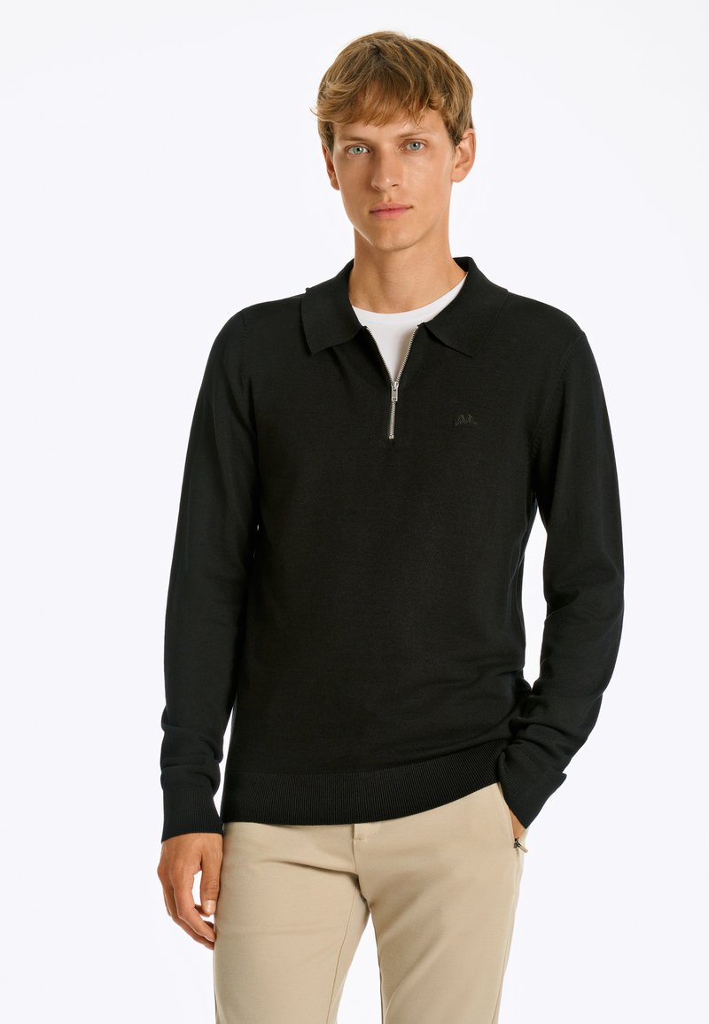 Schwarzer Polo-Pullover mit Reißverschluss und langen Ärmeln, gerippten Säumen und einem weichen Stoff; über einem weißen Hemd getragen. Minimalistisches Logodesign.