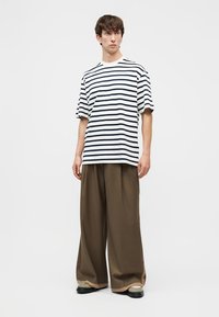 Νέος άνδρας που στέκεται, φοράει λευκό και μαύρο ριγέ κοντομάνικο πουκάμισο, φαρδιά καφέ παντελόνια και μπεζ παπούτσια με μαύρες σόλες.