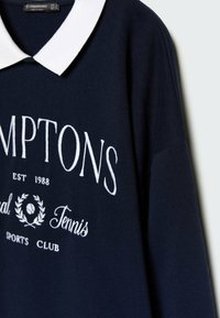 Pull bleu marine avec col blanc, présentant un texte brodé : "HAMPTONS EST 1988" et "Tennis Sports Club" en police audacieuse.