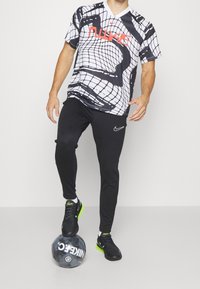 Czarny i biały sportowy strój z siatkowatym wzorem, z czerwonym logo Nike, zestawiony z dopasowanymi czarnymi spodniami i czarnymi butami sportowymi. Piłka nożna znajduje się pod stopą.