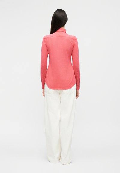 Hosszú, sötét hajú nő, aki testhezálló korallszínű hosszú ujjú felsőt és laza fehér nadrágot visel, és egy fehér háttér felé áll.