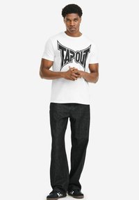 Weißes Baumwoll-T-Shirt mit dem Schriftzug "TAPOUT" in fetten schwarzen Buchstaben, kombiniert mit schwarzen Jeanshosen und schwarzen Sneakers. Einfaches Design, gerade Passform.