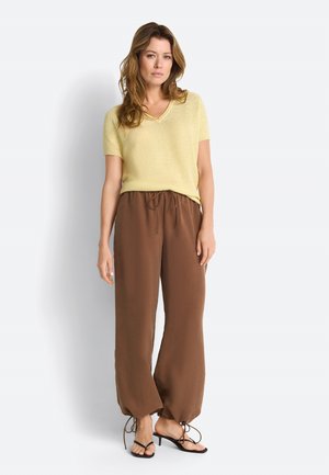 Kvinde iført en lys gul kortærmet top, løse brune bukser med snørebånd ved anklerne og sorte åbne hæle, stående mod en hvid baggrund.
