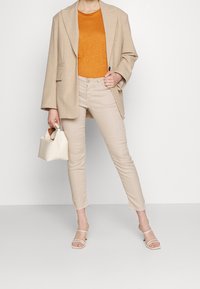 Beige oversize blazer, oranžové tričko, světlé béžové džíny a bílé sandály na podpatku. Malá bílá kabelka s držadlem, hladké textury po celém outfitu.