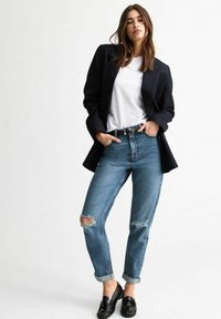 Zwart gestreepte blazer over een wit t-shirt, gecombineerd met lichtblauwe gescheurde jeans en zwarte loafers, weergegeven in een volledige lichaamspositie.