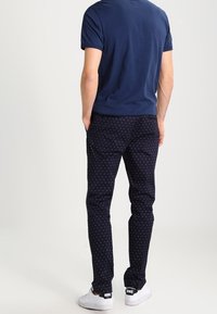 Mężczyzna w granatowej koszulce, granatowych spodniach w małe białe kropki i białych sneakersach Nike, stoi z rękami w kieszeniach, zwrócony tyłem.