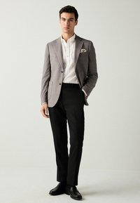 Grau karierter Blazer, weißes Hemd, schwarze maßgeschneiderte Hose und schwarze Schuhe. Der Blazer hat einen einzelnen Knopf und zwei vordere Taschen.
