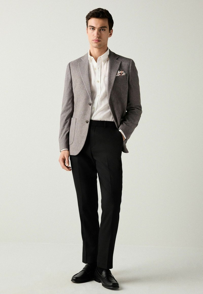 Grau karierter Blazer, weißes Hemd, schwarze maßgeschneiderte Hose und schwarze Schuhe. Der Blazer hat einen einzelnen Knopf und zwei vordere Taschen.
