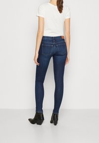 Vaqueros ajustados de denim azul oscuro con cintura alta, que cuentan con dos bolsillos traseros y una etiqueta de cuero, combinados con una camiseta blanca y botines negros.