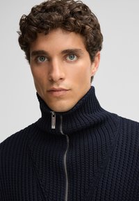 Maglione a collo alto di colore navy con chiusura frontale a zip. Tessuto a coste con un motivo geometrico sul corpo.