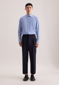 Jasnoniebieska koszula na guziki z wzorem i wąskim kołnierzykiem, zestawiona z granatowymi plisowanymi spodniami i czarnymi sznurowanymi butami.
