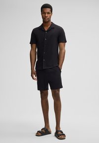 Schwarzes kurzärmliges Hemd mit einem Kragen, Knopfverschluss, kombiniert mit passenden schwarzen Shorts und schwarzen Sandalen mit Riemen und Korksohle.