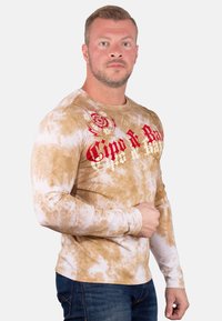 Langarmige, tan kleurige tie-dye shirt met rode grafische tekst en ontwerpen, met een ronde halslijn en een zachte, gladde stoftextuur.