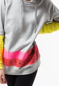 Sweat-shirt gris à capuche, avec des rayures horizontales roses et oranges. Logo noir "adidas" et texte "TELLA SPORT" imprimés en bas.