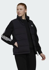 Zwarte gewatteerde vest met hoge kraag, ritssluiting en schouderaccenten. Het materiaal is glad; het heeft een wit Adidas-logo en parallelle strepen.