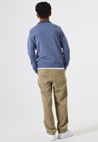 Sudadera azul con cuello redondo, pantalones de pana beige y zapatillas blancas; visto desde atrás sobre un fondo liso.