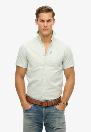 Camisa de botones de manga corta verde claro con cuello y bolsillo en el pecho, combinada con jeans azules y un cinturón de cuero marrón.