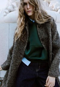 Abrigo de tweed marrón sobre una sudadera verde, con el cuello de una camisa azul visible, combinado con jeans de mezclilla oscuros. El cabello ondulado enmarca el rostro.