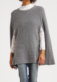 Grå stickad poncho med rund halsringning, sidslitsar och ribbad fåll. Buren över en vit blus med spetskrage, som visar upp lager av texturer.