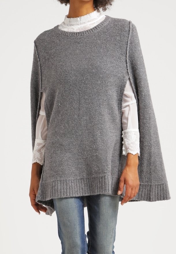 Grå stickad poncho med rund halsringning, sidslitsar och ribbad fåll. Buren över en vit blus med spetskrage, som visar upp lager av texturer.
