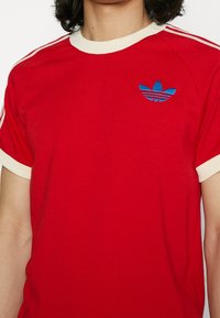 Červené bavlněné tričko s krémovým límečkem a akcenty na rukávech. Obsahuje vyšívané modré a černé logo Adidas na levé části hrudi.