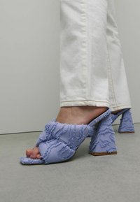 Paarse stoffen muiltjes met gestructureerde franjes, open teen en blokhakken. G gedragen met witte jeans, wat een casual maar gedetailleerd ontwerp laat zien.