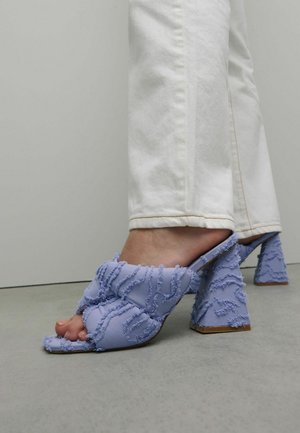 Mules en tissu violet avec des franges texturées, bout ouvert et talons blocs. Portés avec un jean blanc, mettant en valeur un design décontracté mais détaillé.