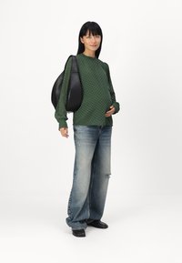 Pull à manches longues texturé de couleur verte avec un col rond, associé à un jean déchiré bleu clair ample et un sac à dos noir arrondi.