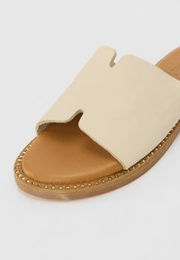 Sandale beige à enfiler avec semelle intérieure couleur fauve, partie supérieure découpée et détails de clous dorés le long du bord de la semelle en bois.