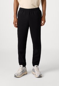 Pantalones deportivos negros con cintura elástica, diseño de pierna recta y tela suave y texturizada. Llevados con zapatillas gris claro que presentan rayas.