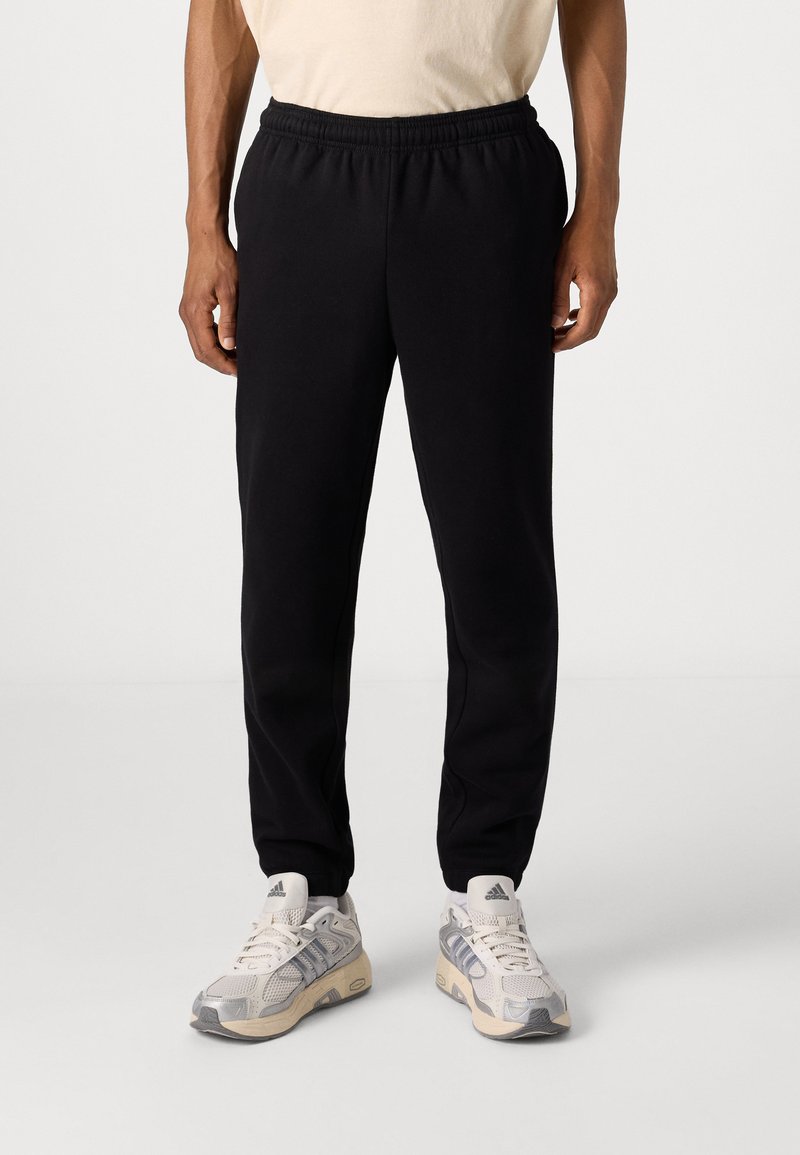 Pantalones deportivos negros con cintura elástica, diseño de pierna recta y tela suave y texturizada. Llevados con zapatillas gris claro que presentan rayas.