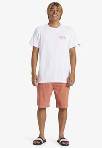 T-shirt en coton blanc avec logo rouge, associé à un short corail et des sandales noires avec des rayures blanches. Tenue décontractée d'été.
