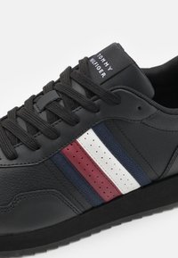 Sneaker in pelle nera con superficie texture, lacci neri e suola in gomma. Strisce notevoli in rosso, bianco e blu navy lungo il lato.