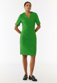 Zielona sukienka z dekoltem w kształcie V, krótkimi rękawami i dopasowanym krojem. Wykonana z gładkiego materiału, sięgająca do kolan. Czarne buty z akcentami.