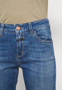 Close-up of blue denim jeans with copper button and yellow stitching, worn with beige fabric top tucked in.