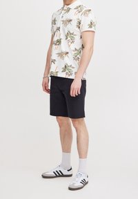 Korte mouwen polo shirt met een crèmekleurige achtergrond en een palmbomenpatroon, gecombineerd met zwarte shorts en witte sneakers met grijze accenten.