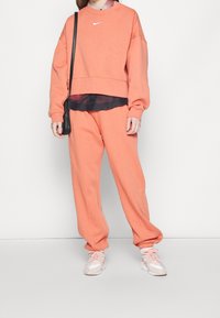 Orange sweatshirt och matchande mjukisbyxor med avslappnad passform, tillverkade av mjukt tyg. Vitt Nike-logga på bröstet. Rosa träningsskor.