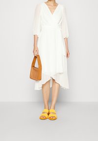 Vit omlottklänning med prickig textur, asymmetrisk fåll och genomskinliga, puffade ärmar. Kombinerad med gula sandaler och en brun handväska.