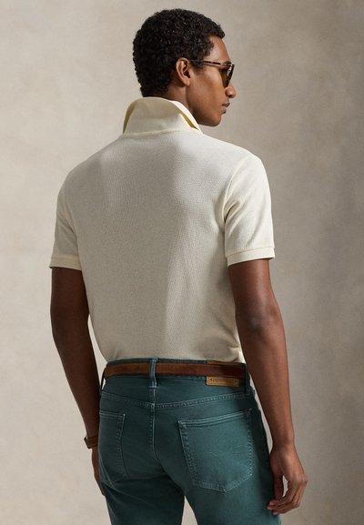 Beige, teksturert polotrøye med krage, brukt med turkise jeans og et brunt lærbelte. Modellen vises bakfra iført solbriller.