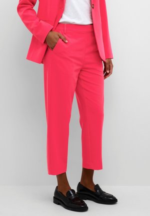Persoon draagt felroze cropped broek en bijpassende blazer, wit overhemd en zwarte loafers, staat met één hand in de zak.