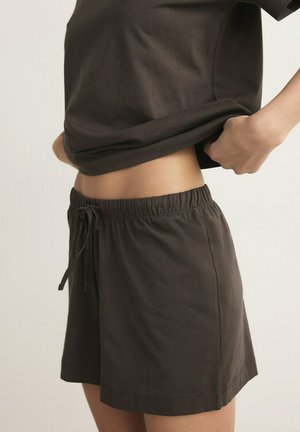 Oseba, oblečena v temno rjave kratke hlače z vrvico, dviguje usklajeno temno rjavo majico, razkrivajoč trebuh na enostavni podlagi.