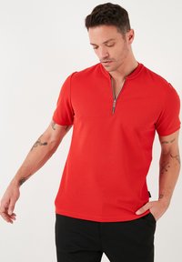 Červené polo tričko s krátkým rukávem a strukturovaným povrchem, s límcem na půl zip. Tričko je v kombinaci s černými kalhotami.