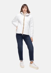 Giacca bianca con cerniera a zip e cappuccio, con cerniera a contrasto gialla e arancione, abbinata a jeans blu scuro con risvolto e sneakers bianche.