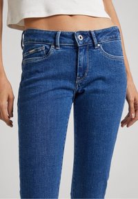 Jeans de mezclilla azul con un diseño ajustado. Estilo tradicional de cinco bolsillos, cierre frontal con botón y detalle de costura visible en la cintura.