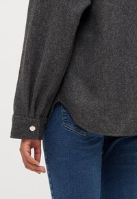 Szara flanelowa koszula z długimi rękawami i zapięciem na guzik, z zaokrąglonym dołem. Noszona z niebieskimi dżinsami, z widocznym detalem tekstury materiału.