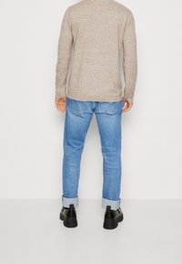 Man som bär en beige långärmad tröja, ljusblå uppvikt jeans och svarta skor, stående mot en enkel vit bakgrund med ryggen vänd mot kameran.