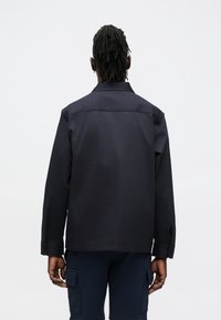 Camisa azul marino con cuello y mangas largas. Presenta un corte recto y puños con botones, confeccionada en tela lisa. Detalles de costuras visibles.