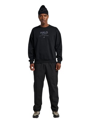 Hombre con gorro negro, sudadera negra con texto "HALO", pantalones negros y zapatillas grises, de pie sobre fondo blanco.