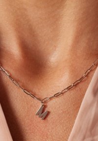 Zilveren ketting met ovale schakels, met een metalen hanger in de vorm van de letter "M". Close-up met nadruk op de zachte textuur en de gepolijste afwerking.